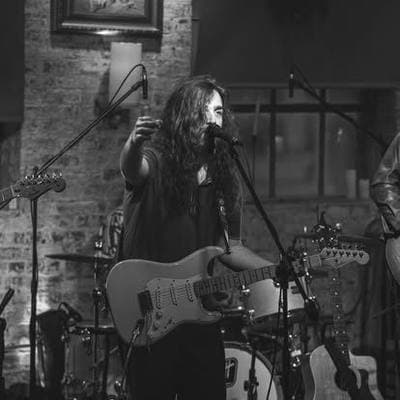 Black and white: a long-haired guitarist playing in front of exposed brick.