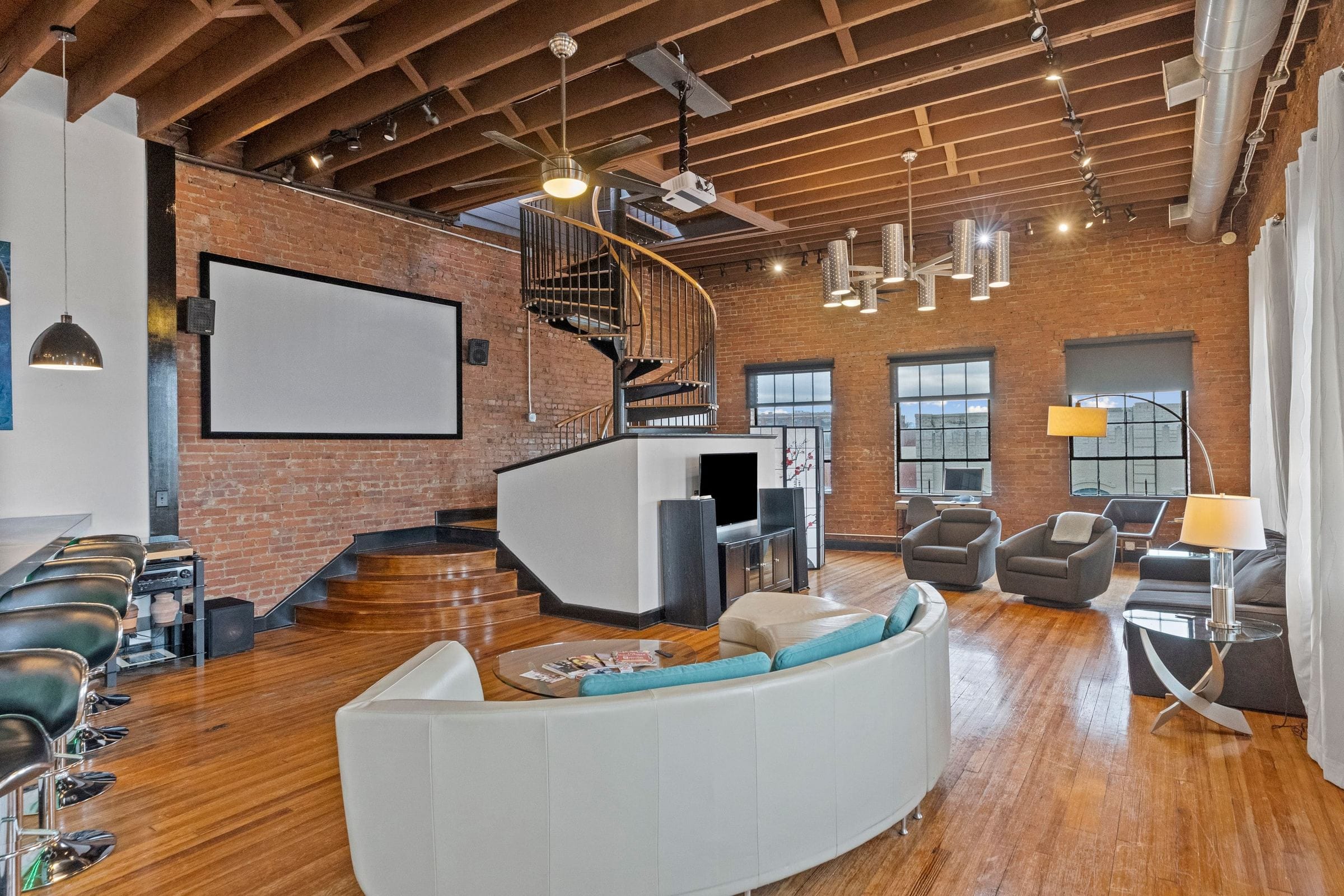 The black spiral stair rising from a raised platform beside a projector screen, the curved sofa in front.