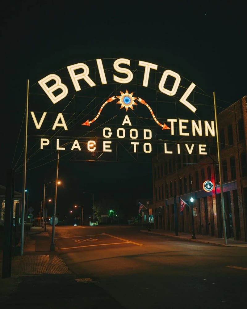 The illuminated Bristol sign arching over State Street at night, reading VA and TENN on either side of A Good Place to Live, with downtown Bristol's brick buildings beside the empty street.