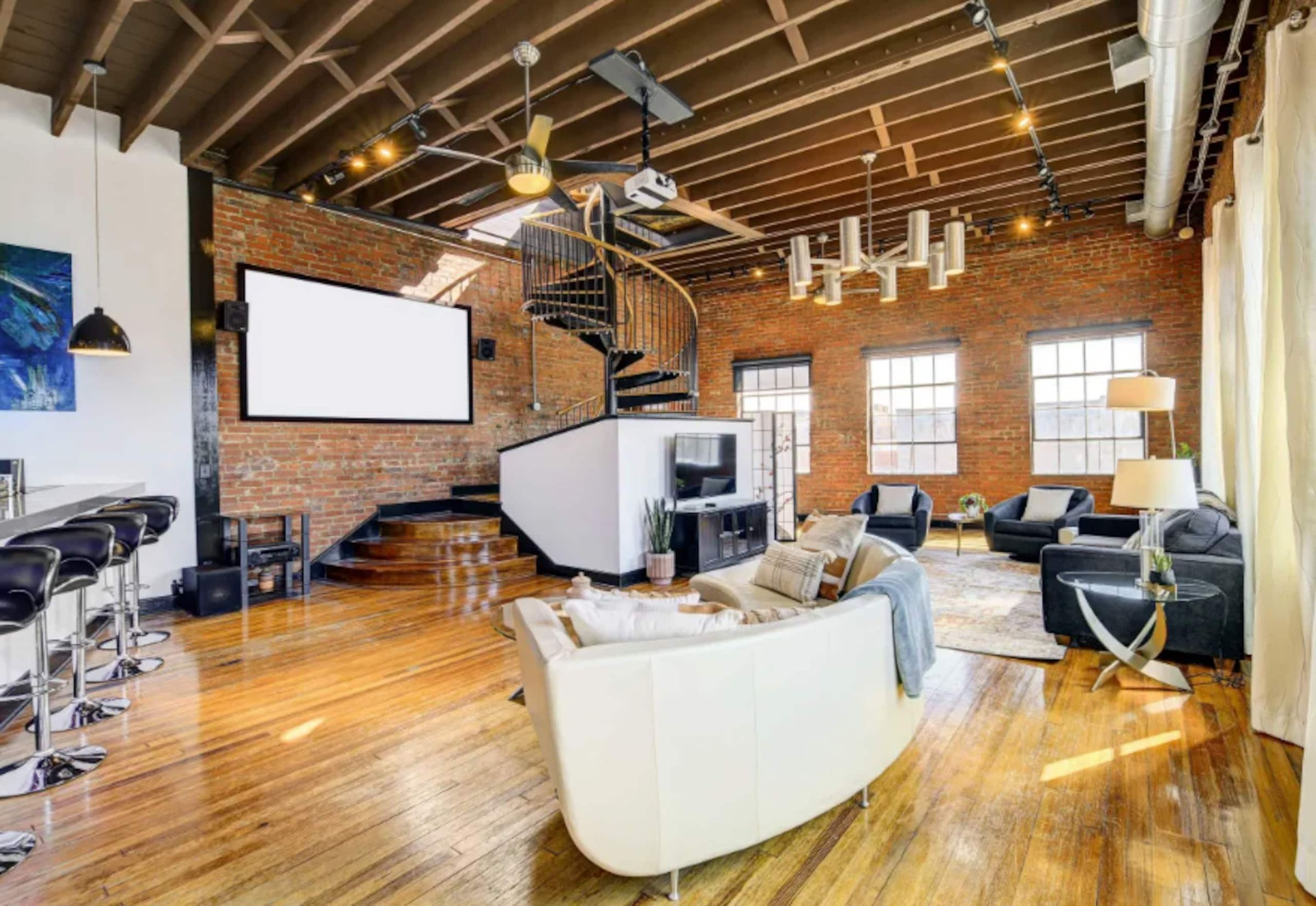 The loft's open main floor: exposed brick and timber beams, tall windows, the black spiral stair at the back, and a curved white sofa.