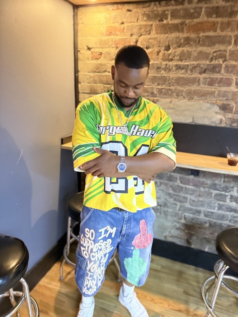 DJ Pharoh in a green and yellow jersey and hand-painted denim shorts, arms folded, in front of exposed brick.